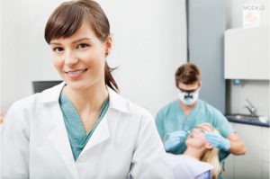 Female Dentist Smiling at Camera with Assistant and Patient in Background