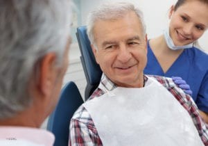 Mature Male In Plaid Shirt Sitting In Dental Chair Talking To Doctor With Assistant Smiling
